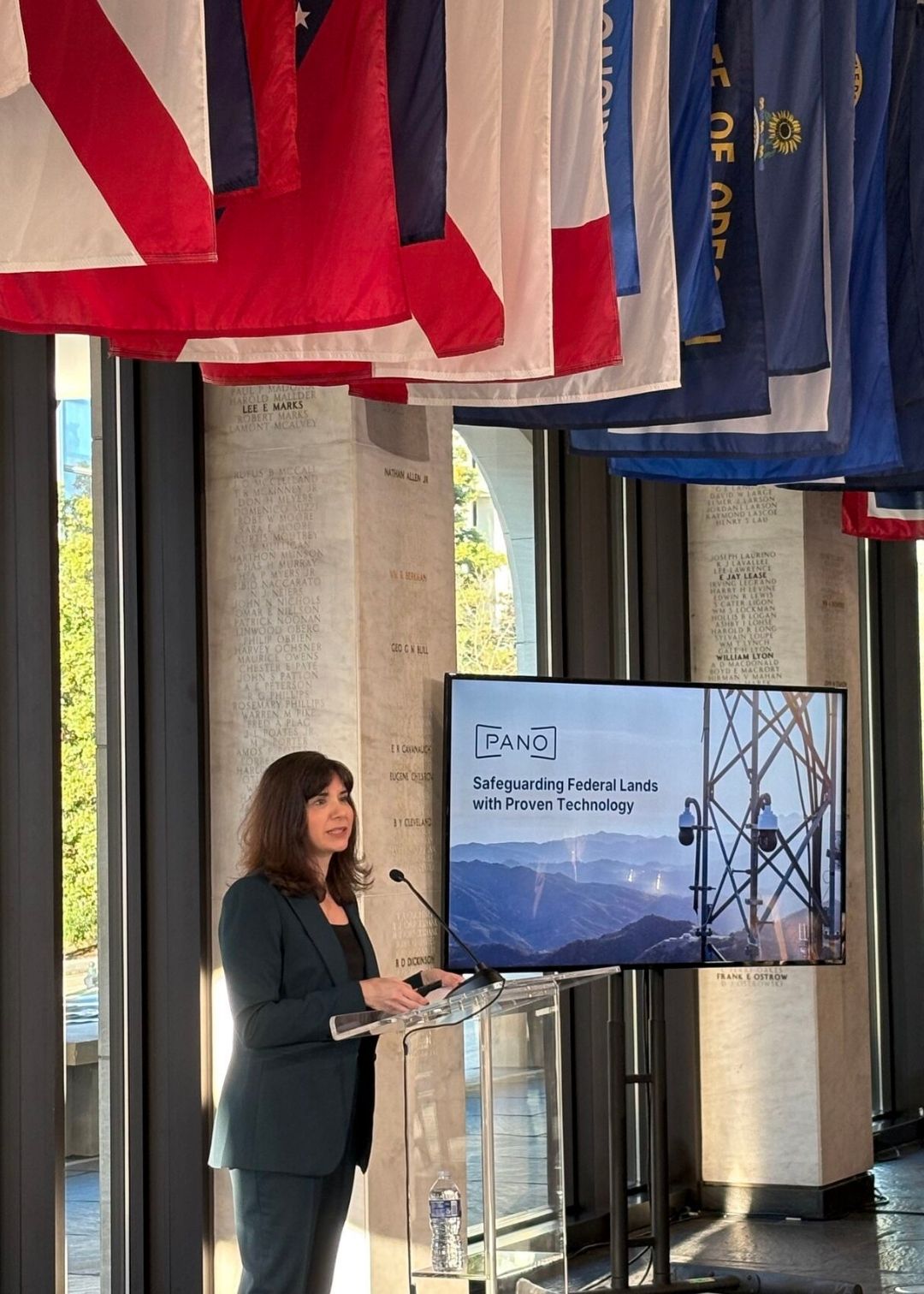 A woman in a suit speaks at a podium next to a screen displaying "PANO: Safeguarding Federal Lands with Proven Technology and Testimonials." Various flags hang from the ceiling above.